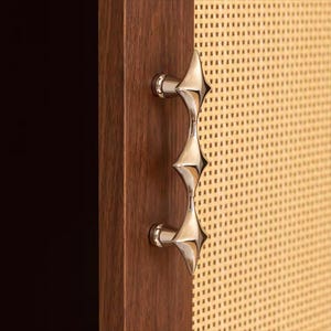 以下が含まれることがあります： ユニークなスターバーストデザインの、磨かれたシルバーのキャビネットプル。ハンドルは、織り込まれた明るい色のパネルが付いた、濃い色の木製キャビネットドアに取り付けられています。この金具は、モダンなエレガンスを添えています。