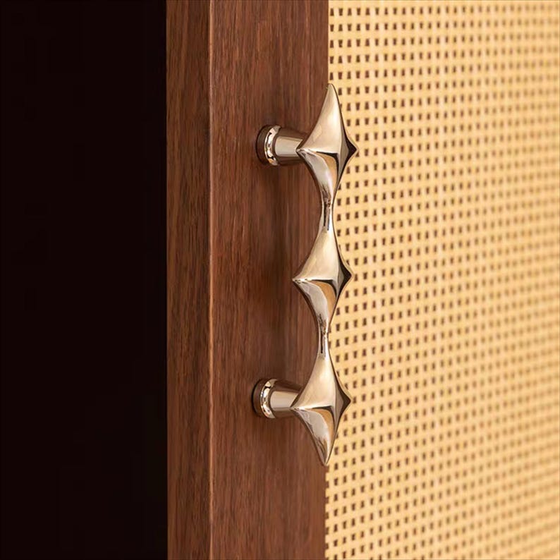 以下が含まれることがあります： ユニークなスターバーストデザインの、磨かれたシルバーのキャビネットプル。ハンドルは、織り込まれた明るい色のパネルが付いた、濃い色の木製キャビネットドアに取り付けられています。この金具は、モダンなエレガンスを添えています。