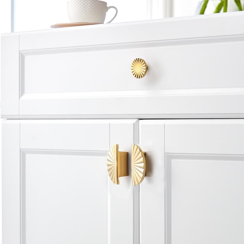 May include: Close-up of white cabinet doors and drawer with gold-coloured hardware. The drawer features a round, sunburst-style knob. The cabinet doors have half-circle handles with a fan-like design. A white teacup sits on top of the cabinet.