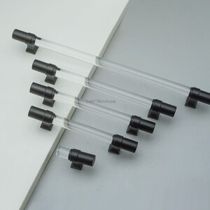 K&ouml;nnte beinhalten: Eine Sammlung von Schrankgriffen in verschiedenen Gr&ouml;&szlig;en. Jeder Griff hat einen transparenten, zylindrischen K&ouml;rper mit schwarzen Endkappen und Befestigungen. Pr&auml;sentiert auf einer hellgrauen und wei&szlig;en Oberfl&auml;che.