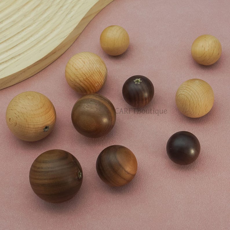 May include: A collection of wooden spheres in various shades of brown and tan, arranged on a pink surface. The spheres vary in size and wood grain patterns, with some featuring small drilled holes. The overall aesthetic is natural and rustic.
