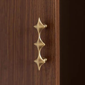 以下が含まれることがあります： 真鍮色のスターバーストデザインのキャビネットプル。ハンドルは濃い茶色の木製キャビネットドアに取り付けられています。プルは、中央のステムで接続された3つのダイヤモンド型の要素を備え、装飾的で機能的なハードウェアピースを作成します。