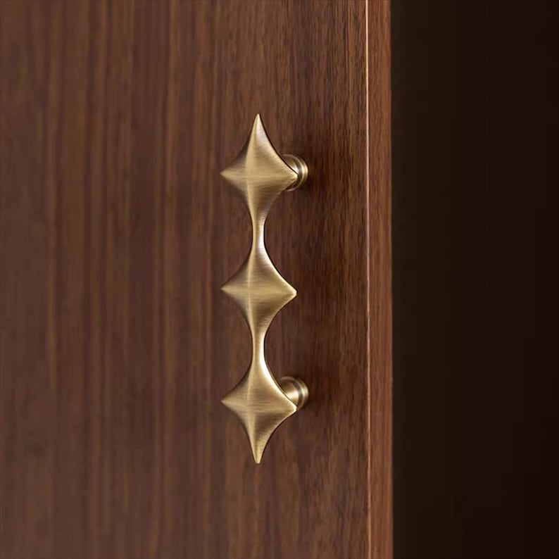 以下が含まれることがあります： 真鍮色のスターバーストデザインのキャビネットプル。ハンドルは濃い茶色の木製キャビネットドアに取り付けられています。プルは、中央のステムで接続された3つのダイヤモンド型の要素を備え、装飾的で機能的なハードウェアピースを作成します。