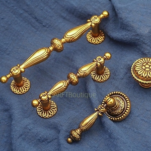 May include: Ornate gold-colored cabinet hardware set, including two handles and two knobs. The handles have a curved design with decorative detailing. The knobs feature a floral pattern. The hardware is set against a blue background.