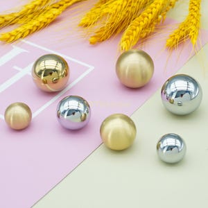 May include: A collection of metallic spheres in different sizes and finishes, including gold and silver, are displayed on a two-tone pink and cream surface. The spheres reflect light, creating highlights. Yellow wheat stalks are in the background.