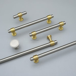 K&ouml;nnte beinhalten: Eine Auswahl an Schrankgriffen und -kn&ouml;pfen in geb&uuml;rsteter Silberausf&uuml;hrung mit goldfarbenen Details. Das Bild zeigt eine Vielzahl von Gr&ouml;&szlig;en und Stilen, darunter ein runder Knopf und Stangengriffe in verschiedenen L&auml;ngen.