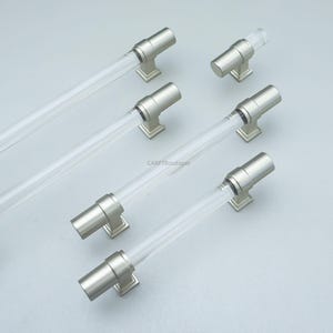 K&ouml;nnte beinhalten: Schrankgriffe mit klarem, zylindrischem Mittelteil und Enden und Sockeln aus geb&uuml;rstetem, nickelfarbenem Metall. Die Griffe sind diagonal auf einer hellgrauen Oberfl&auml;che angeordnet. Der Text "CARFTBoutique" ist sichtbar.