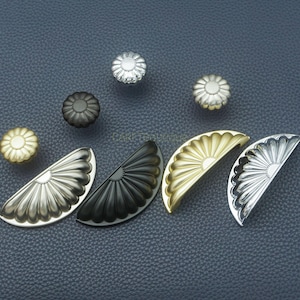 May include: A collection of decorative cabinet hardware, including flower-shaped knobs and half-circle pulls. The knobs are in silver, gold, and black finishes. The pulls are in silver, gold, and black finishes. The hardware is displayed on a dark blue surface.