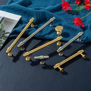 K&ouml;nnte beinhalten: Eine Auswahl an Schrankgriffen und einem Knopf in Gold- und Silberausf&uuml;hrungen. Das Bild zeigt verschiedene Griffdesigns, jeweils mit dekorativen Basen. Die Artikel sind auf einem blauen Stoff angeordnet, mit roten Blumen im Hintergrund.