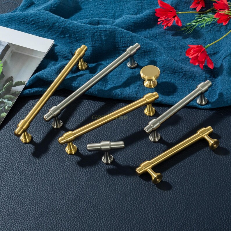 K&ouml;nnte beinhalten: Eine Auswahl an Schrankgriffen und einem Knopf in Gold- und Silberausf&uuml;hrungen. Das Bild zeigt verschiedene Griffdesigns, jeweils mit dekorativen Basen. Die Artikel sind auf einem blauen Stoff angeordnet, mit roten Blumen im Hintergrund.