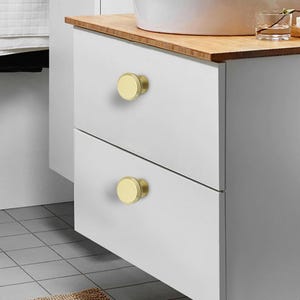 Pode incluir: Close-up de uma bancada de casa de banho branca com duas gavetas, cada uma com um puxador redondo dourado. Os puxadores t&ecirc;m um design simples e moderno. A bancada &eacute; encimada por uma superf&iacute;cie de madeira clara, e uma pia branca &eacute; vis&iacute;vel.