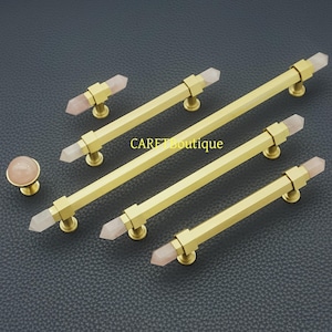 May include: A collection of gold-coloured cabinet hardware, including handles and a knob. The handles have a rectangular design with a light pink crystal at each end. The knob is round with a matching pink crystal. The text "CARET boutique" is visible.