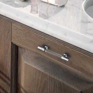 K&ouml;nnte beinhalten: Ein klarer Acryl- und Chrom-Schrankgriff ist an einer h&ouml;lzernen Schrankt&uuml;r montiert. Der Griff hat eine zylindrische Acrylmitte und Chromendkappen. Die Schrankt&uuml;r hat eine nat&uuml;rliche Holzmaserung.