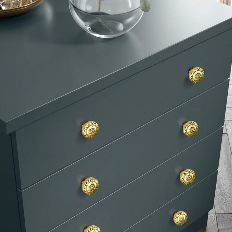 May include: A dark teal dresser with five drawers, each adorned with a gold-colored, ornate knob. The knobs have a detailed, circular design. A glass vase and decorative bowl sit on top of the dresser.