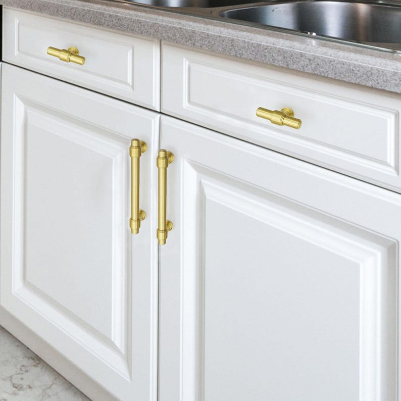 Pode incluir: Arm&aacute;rios de cozinha brancos com ferragens douradas. A imagem mostra dois arm&aacute;rios com uma bancada e uma pia ao fundo. As portas dos arm&aacute;rios t&ecirc;m puxadores e puxadores dourados.