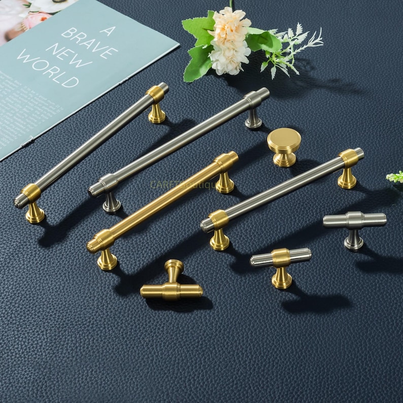 K&ouml;nnte beinhalten: Eine Auswahl an Schrankgriffen und -kn&ouml;pfen in Gold- und Silberausf&uuml;hrungen. Die Hardware ist auf einer dunkelblauen Oberfl&auml;che angeordnet, mit einer Zeitschrift mit dem Text "A Brave New World" im Hintergrund.