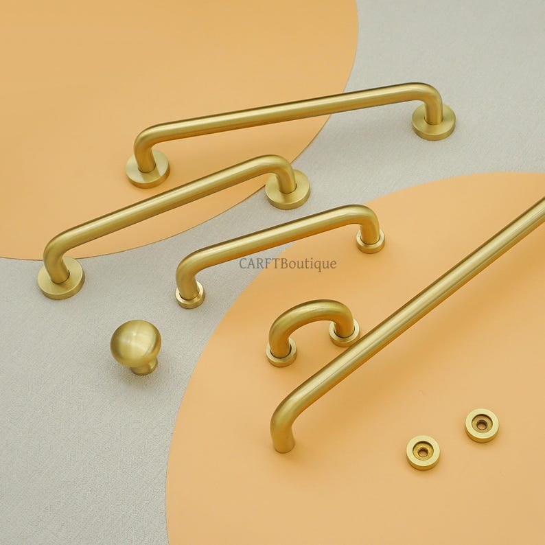 May include: A selection of gold-toned cabinet handles and a knob, displayed on a neutral and peach-colored background. The set includes several bar pulls of different lengths, along with two small circular fixings.