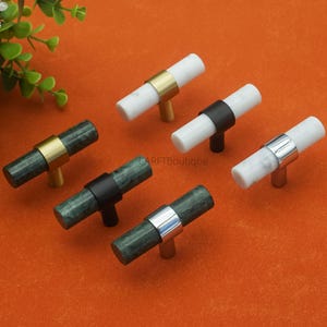 May include: Six cabinet knobs with a T-bar design. The knobs feature cylindrical marble bars in white and green, accented with gold, silver, and black metal rings and stems. The knobs are arranged on an orange surface.