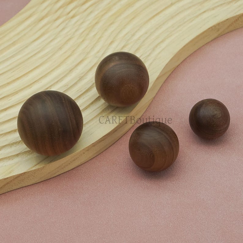 May include: Four wooden spheres of varying sizes, arranged on a light wood surface with a pink background. The spheres are a rich brown color, with visible wood grain. The text "CARETBoutique" is visible on the wood.