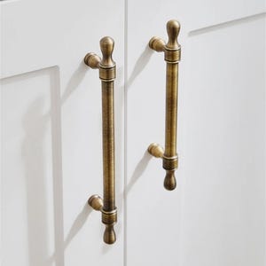 K&ouml;nnte beinhalten: Nahaufnahme von zwei antiken Messing-Schrankgriffen, die an einer wei&szlig;en Schrankt&uuml;r befestigt sind. Die Griffe haben ein vertikales Stangendesign mit dekorativen Endst&uuml;cken und verleihen der Schrankwand einen Vintage-Touch.