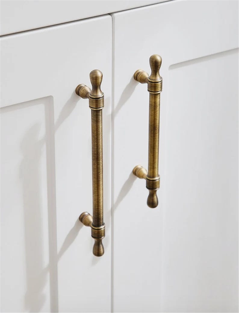 K&ouml;nnte beinhalten: Nahaufnahme von zwei antiken Messing-Schrankgriffen, die an einer wei&szlig;en Schrankt&uuml;r befestigt sind. Die Griffe haben ein vertikales Stangendesign mit dekorativen Endst&uuml;cken und verleihen der Schrankwand einen Vintage-Touch.