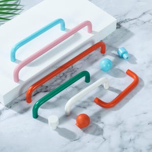 May include: A collection of colorful cabinet hardware, including blue, pink, red, green, and white handles. Also shown are matching knobs and pulls in various shapes and colors. The hardware is arranged on a white surface against a marble background.