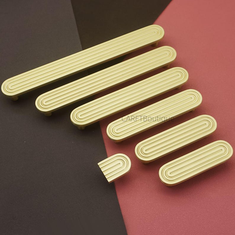 Puede incluir: Un conjunto de tiradores dorados para armarios con un dise&ntilde;o acanalado. Los tiradores est&aacute;n dispuestos sobre un fondo bicolor burdeos y marr&oacute;n oscuro. El texto "CARETBoutique" es visible en el fondo.