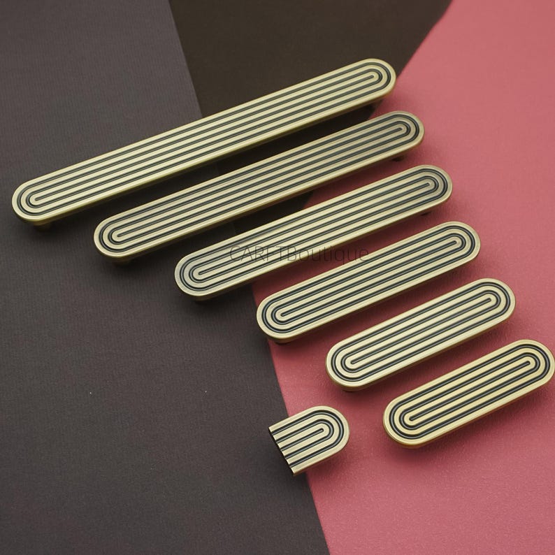 Puede incluir: Colecci&oacute;n de tiradores de armario de lat&oacute;n antiguo en varios tama&ntilde;os. Cada tirador presenta una serie de l&iacute;neas paralelas y ranuradas dentro de una forma ovalada. El herraje se muestra sobre un fondo bicolor marr&oacute;n y rosa.