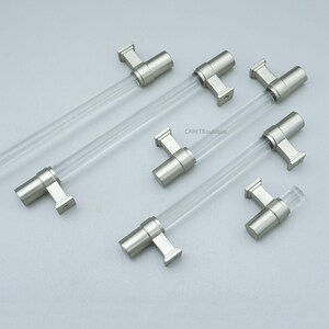 K&ouml;nnte beinhalten: Sechs Schrankgriffe mit modernem Design. Jeder Griff hat einen transparenten, zylindrischen Griff und eine Halterung in geb&uuml;rstetem Nickel. Die Griffe sind auf einer hellgrauen Oberfl&auml;che angeordnet.