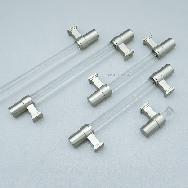 K&ouml;nnte beinhalten: Sechs Schrankgriffe mit modernem Design. Jeder Griff hat einen transparenten, zylindrischen Griff und eine Halterung in geb&uuml;rstetem Nickel. Die Griffe sind auf einer hellgrauen Oberfl&auml;che angeordnet.