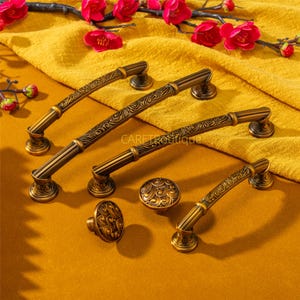 Könnte beinhalten: Antikes bronzefarbenes Schrankbeschlagsset. Das Set enthält vier Griffe und zwei Knöpfe mit verzierten floralen Designs. Die Griffe sind gebogen und mit dekorativen Details versehen. Die Hardware wird auf einer gelben Oberfläche präsentiert.