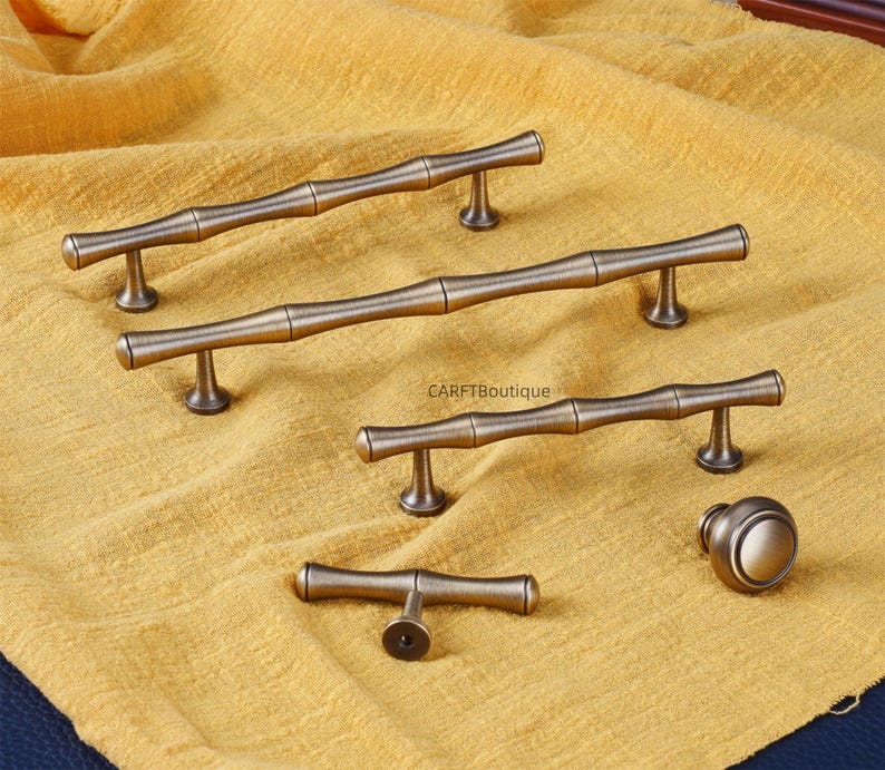 K&ouml;nnte beinhalten: Eine Sammlung von antiken bronzefarbenen Schrankbeschl&auml;gen, darunter mehrere Griffe im Bambus-Stil und ein runder Knopf. Die Beschl&auml;ge sind auf einem strukturierten gelben Stoff ausgestellt. Der Text "CARFTBoutique" ist sichtbar.