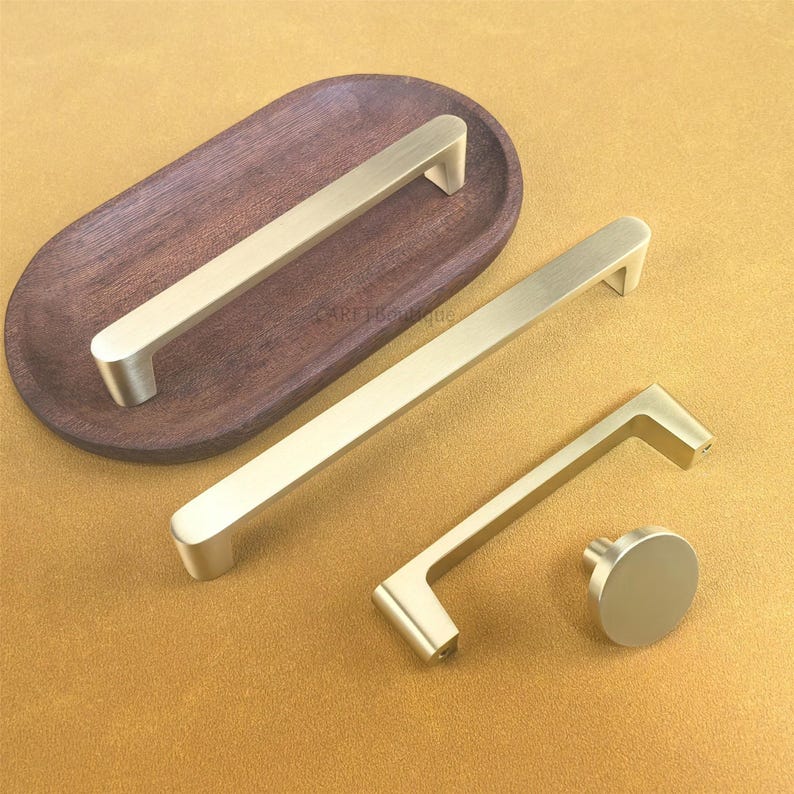 May include: A selection of gold-coloured cabinet hardware, including two long bar pulls, a smaller pull, and a round knob. One bar pull rests on a dark brown wooden tray. The hardware is displayed on a mustard-coloured surface.