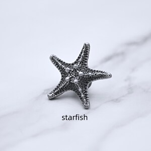 Peut inclure: Un objet en forme d'&eacute;toile de mer de couleur argent&eacute;e avec des d&eacute;tails textur&eacute;s et une petite &eacute;pingle au dos. Le mot "starfish" est imprim&eacute; en dessous. Il s'agit probablement d'une broche ou d'une &eacute;pingle.