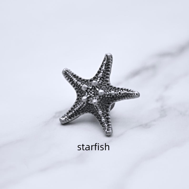 Peut inclure: Un objet en forme d'&eacute;toile de mer de couleur argent&eacute;e avec des d&eacute;tails textur&eacute;s et une petite &eacute;pingle au dos. Le mot "starfish" est imprim&eacute; en dessous. Il s'agit probablement d'une broche ou d'une &eacute;pingle.