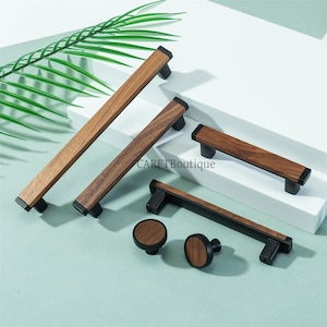 May include: A collection of wooden cabinet pulls and knobs. The pulls have a rectangular wooden center with black ends and bases. The knobs are round with a wooden face and black base. The text "CARETBoutique" is visible.