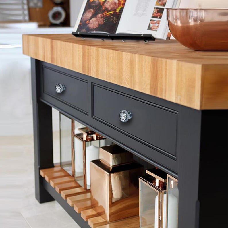 May include: A kitchen island with a butcher block countertop and black cabinetry. The island features two drawers with crystal knobs, open shelving, and decorative copper-toned accents. A cookbook and copper bowl are on the countertop.