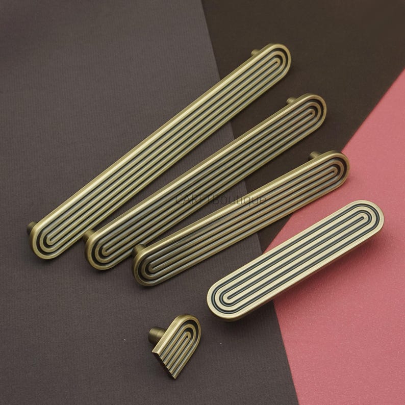 May include: A collection of antique brass cabinet hardware, including several elongated pulls and a small knob. The hardware features a grooved design with a dark accent, set against a brown and pink background.