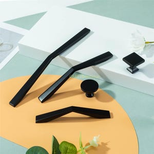 May include: A collection of black cabinet hardware, including handles and knobs. The handles have a sleek, modern design, with a variety of shapes and sizes. The knobs include a square and a round design. The hardware is displayed on a white and yellow surface.