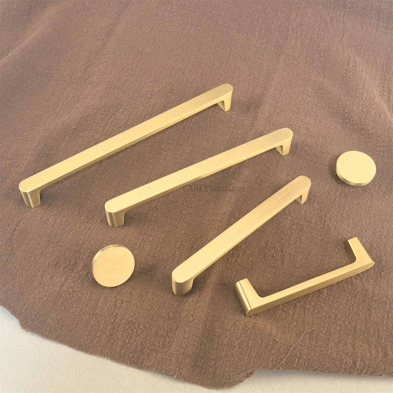 May include: A selection of gold-coloured cabinet handles and knobs. The image shows several rectangular bar pulls and two circular knobs. The hardware is displayed on a brown fabric, highlighting the different sizes and styles for home projects.