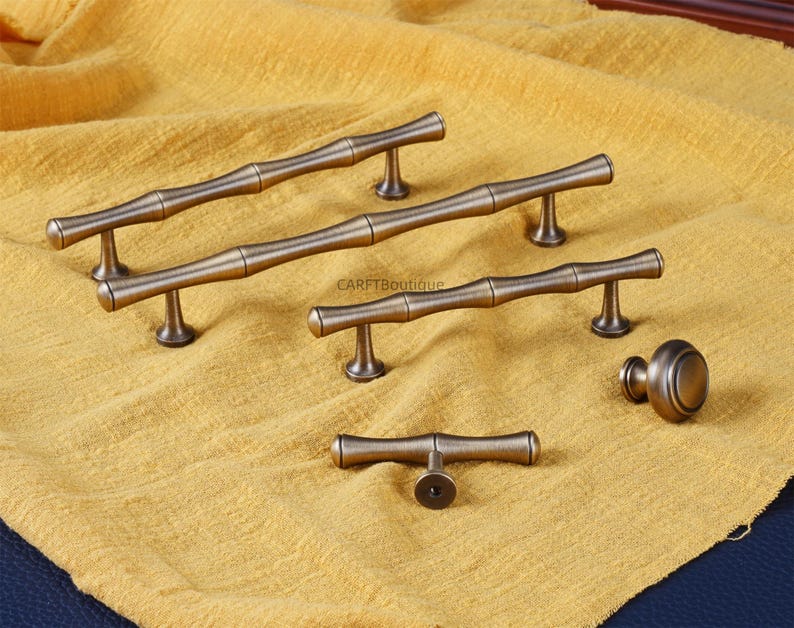 K&ouml;nnte beinhalten: Eine Sammlung von M&ouml;belbeschl&auml;gen in Bronzefarbe, darunter mehrere Griffe im Bambus-Stil und ein runder Knauf. Die Beschl&auml;ge sind auf einem strukturierten gelben Stoff angeordnet, um das Design und die Oberfl&auml;che zu zeigen.