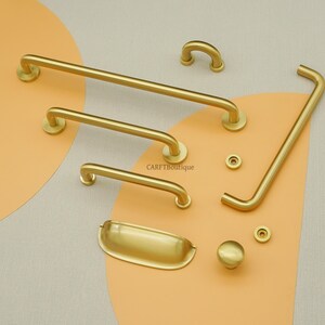 May include: A selection of gold-coloured cabinet hardware, including handles, knobs, and a pull. The hardware has a modern, minimalist design with rounded edges and a matte finish. The pieces are arranged on a neutral background with an orange accent.