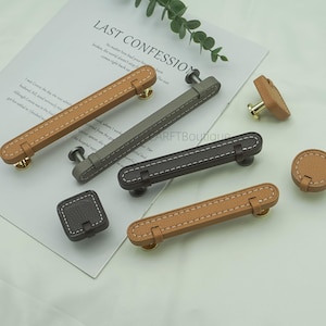 May include: A collection of cabinet hardware, including handles and knobs, in various shades of brown, gray, and black. The hardware features a leather-like texture with white stitching and metal accents. The text "LAST CONFESSION" is visible in the background.