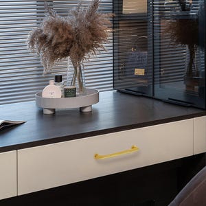 May include: A contemporary dressing table with a dark work surface and white drawer fronts. A gold-coloured handle is fixed to the drawer. A decorative tray with a vase of dried flowers and perfume bottles sits on the surface. A glass cabinet is in the background.