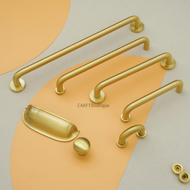 May include: A selection of gold-toned cabinet hardware, featuring bar pulls of different lengths, a cup pull, a round knob, and small circular accents. The items are displayed on a peach-coloured surface against a neutral backdrop. The brand name "CARFTBoutique" is visible.