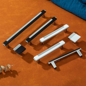 Può includere: Un assortimento di maniglie per mobili e un pomello quadrato, in bianco e nero con dettagli argentati. Le maniglie sono esposte su una superficie arancione. Misure in centimetri.