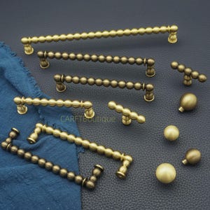 Puede incluir: Una colección de herrajes de latón antiguo para gabinetes, que incluye manijas y perillas de varios tamaños. Las manijas presentan un diseño de cuentas y las perillas son esféricas. Los herrajes se exhiben sobre una superficie oscura con un paño azul.