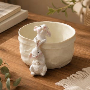 Puede incluir: Un cuenco de cerámica de color crema con un acabado texturizado similar al papel. Dos pequeñas figuras de conejos blancos están unidas al borde frontal, con un conejo de pie dentro del cuenco y el otro colgando debajo, ambos con sutiles acentos rosados.