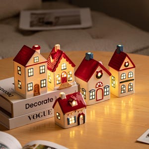 Puede incluir: Cinco figuras de casas en miniatura de cerámica con interiores iluminados, con techos rojos y paredes blancas. Las casas tienen varios detalles arquitectónicos como ventanas, puertas y coronas, y están dispuestas sobre libros apilados.