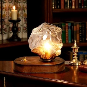 Könnte beinhalten: Eine dekorative Tischlampe mit einem strukturierten Glasschirm, der ein warmes, goldenes Leuchten ausstrahlt. Die Lampe steht auf einem rechteckigen Holzsockel und verleiht jedem Raum einen Hauch von Eleganz. Ein stilvolles Wohnaccessoire.